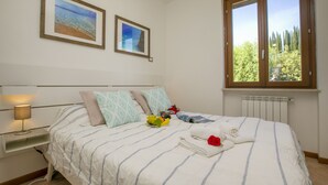 Ein ordentlich bezogenes Bett mit Streifenmuster, ein Nachttisch mit Lampe, zwei gerahmte Bilder an der Wand und ein Fenster mit Blick auf Bäume.