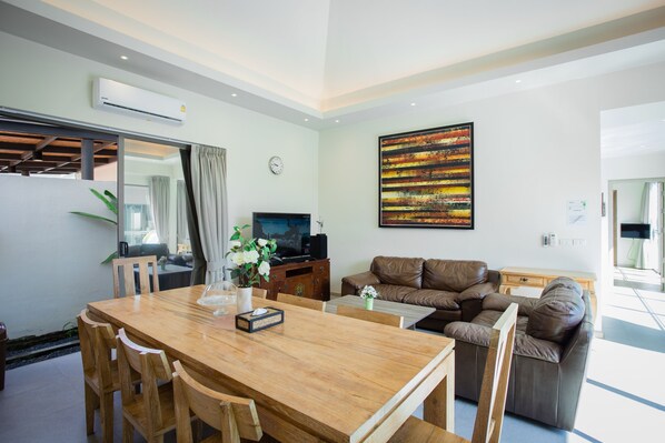 A modern living room with a wooden dining table, brown leather sofa, and a large abstract painting on the wall. - Yipmunta Pool Villa (Choeng Thale)