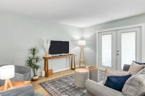 A modern living room with a flat-screen TV, a wooden TV stand, a floor lamp, a grey sofa, a patterned rug, and a potted plant. - Entire Beach House, Steps to Beach, Heated Pool, Balcony, Walk to Restaurants, $300 in Free Tickets! (St. Augustine)