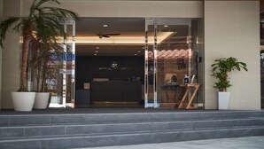 Entrada de hotel com balcão de recepção, palmeiras e escadaria.