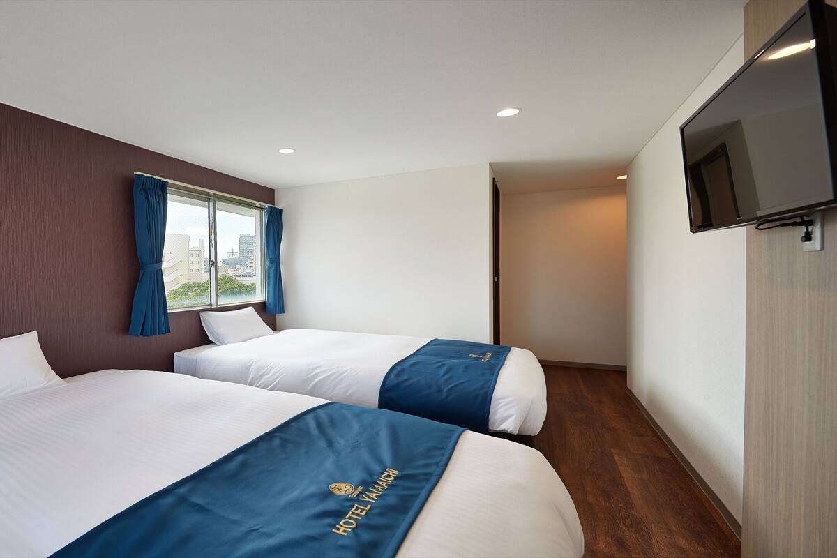 A hotel room with two beds, a large window with curtains, a flat-screen TV mounted on the wall, and a wooden floor.