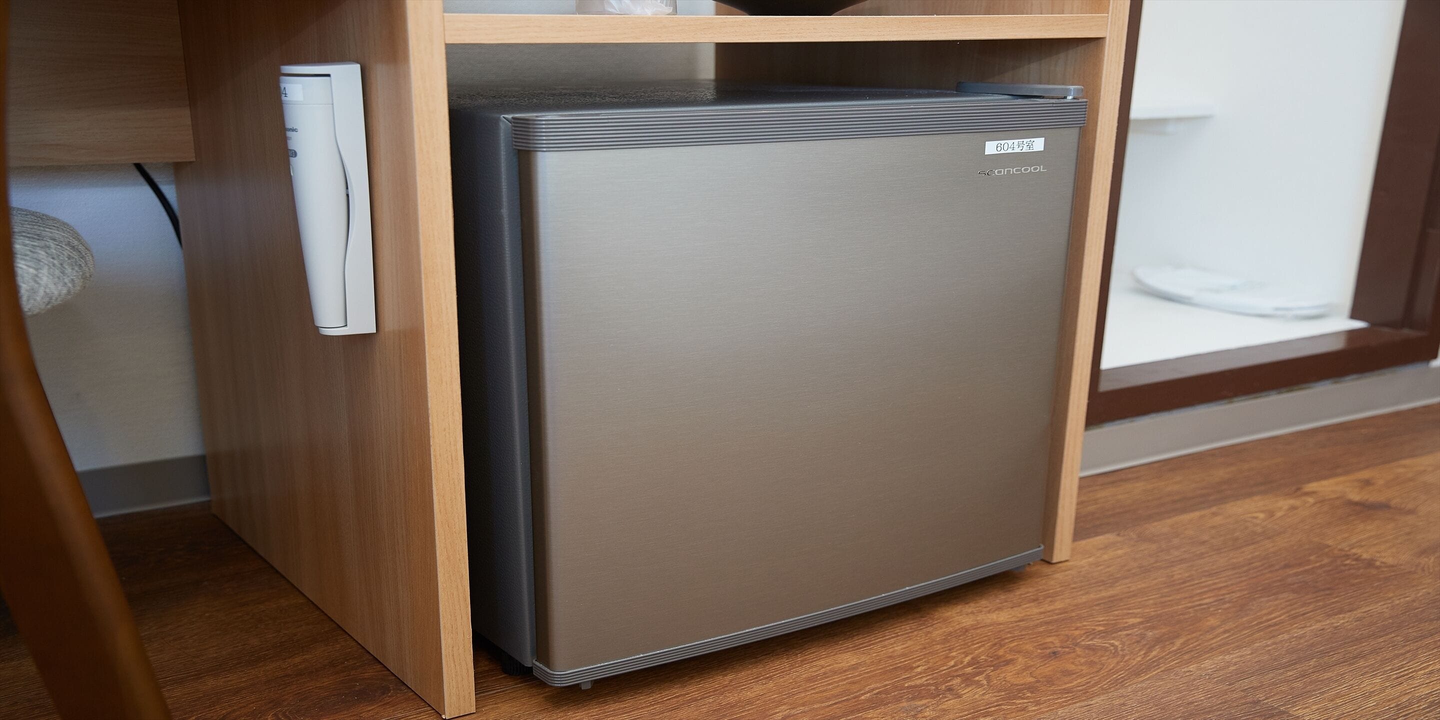 A compact, stainless steel mini-fridge with a digital display, placed under a wooden shelf in a room with hardwood flooring.