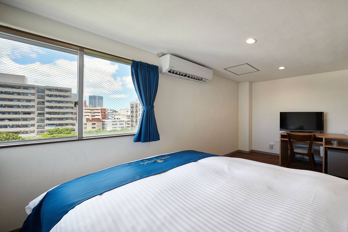 A hotel room with a bed, a desk with a television, a small refrigerator, and a window with a curtain.
