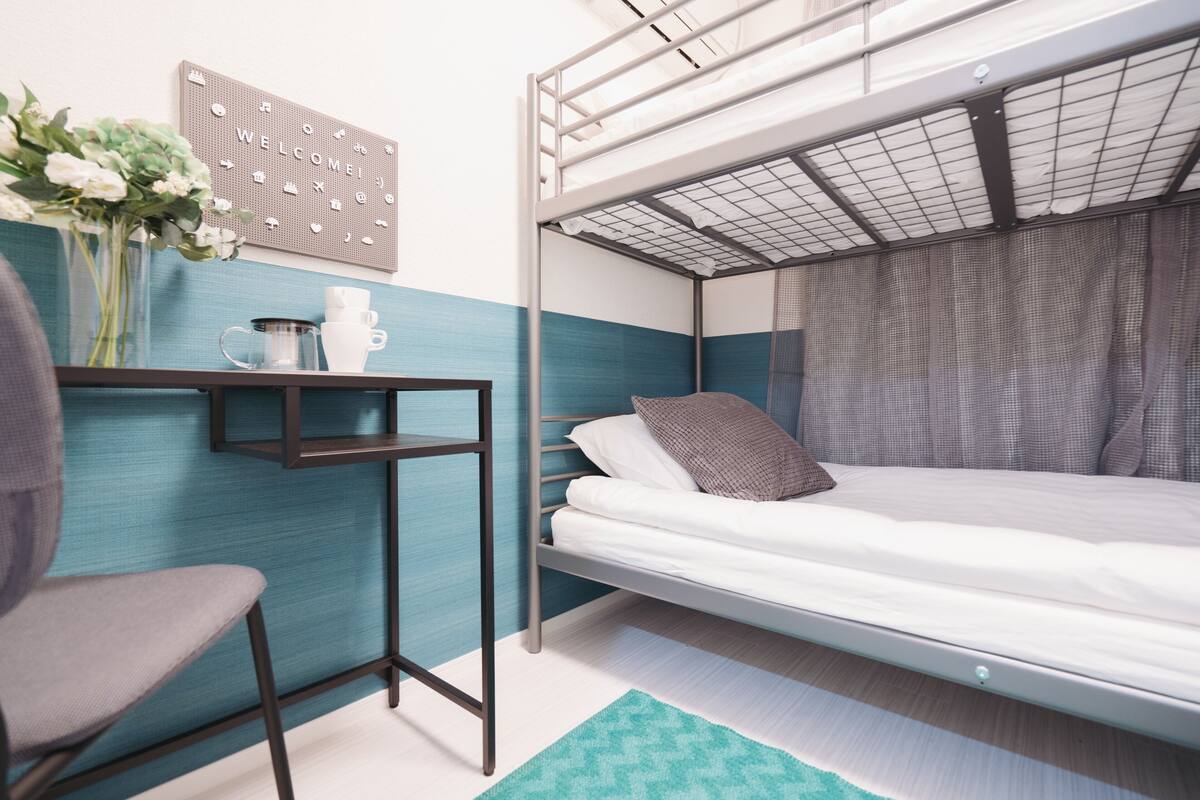 A bunk bed with a metal frame, a small desk with a chair, a coffee maker, and a welcome sign on the wall.