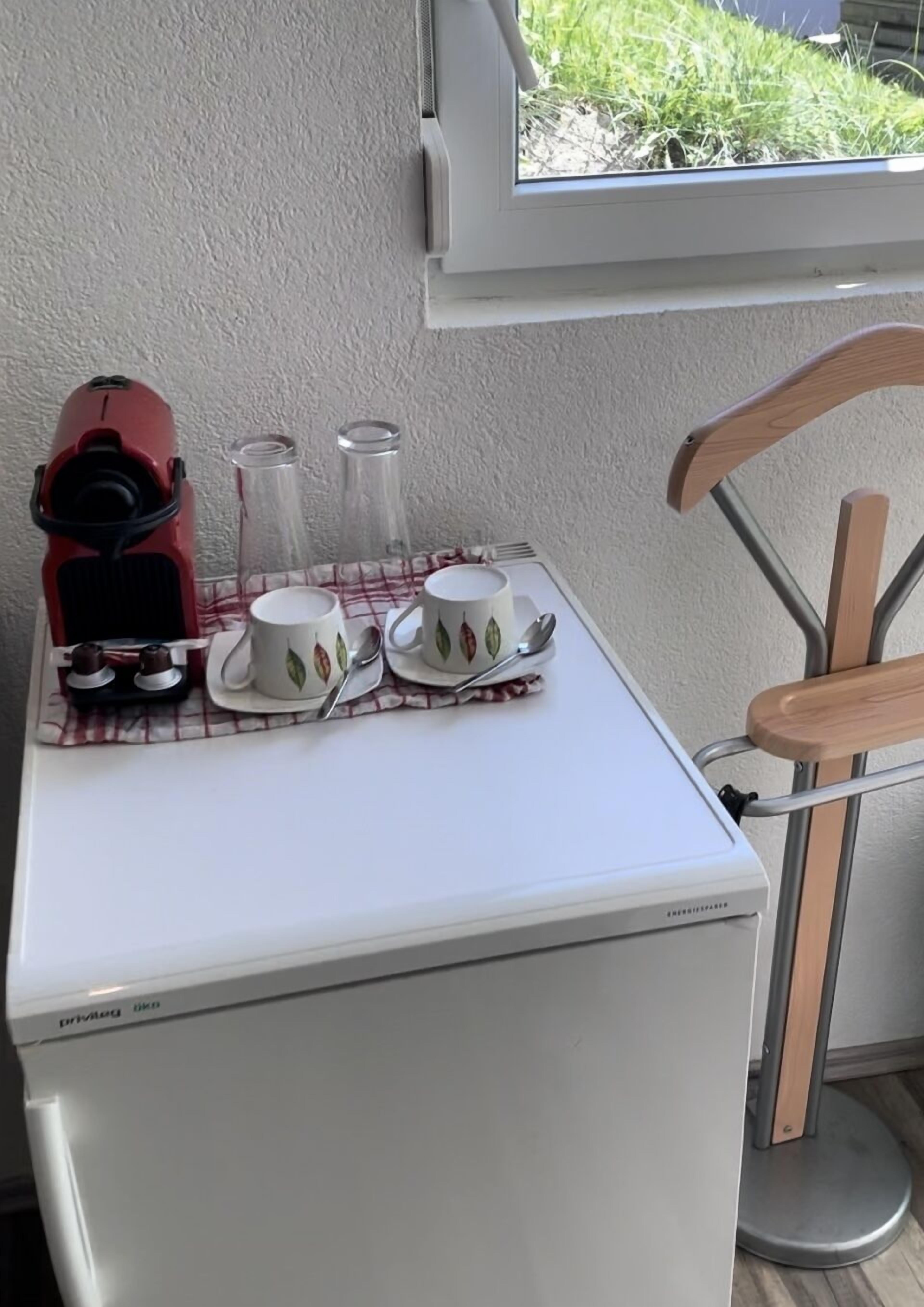 Una kitchenette compacta con un refrigerador blanco, una cafetera roja, dos tazas con diseños de hojas y una silla de metal.
