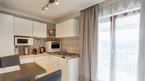 Een moderne keuken met witte kastjes, een magnetron, een oven en een spoelbak. Er is een eettafel met stoelen en een groot raam met gordijnen.
