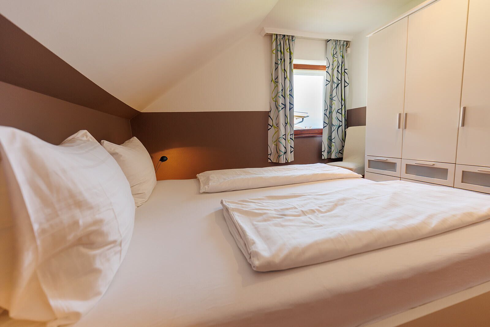 Een kamer met een enkel bed, een bruine accentmuur, wit beddengoed en een raam met geruit gordijnen.