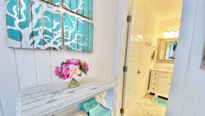 Un pasillo con una puerta blanca, un espejo y un tocador con lavamanos. Se observan paneles decorativos en la pared y un florero con flores sobre una estantería.