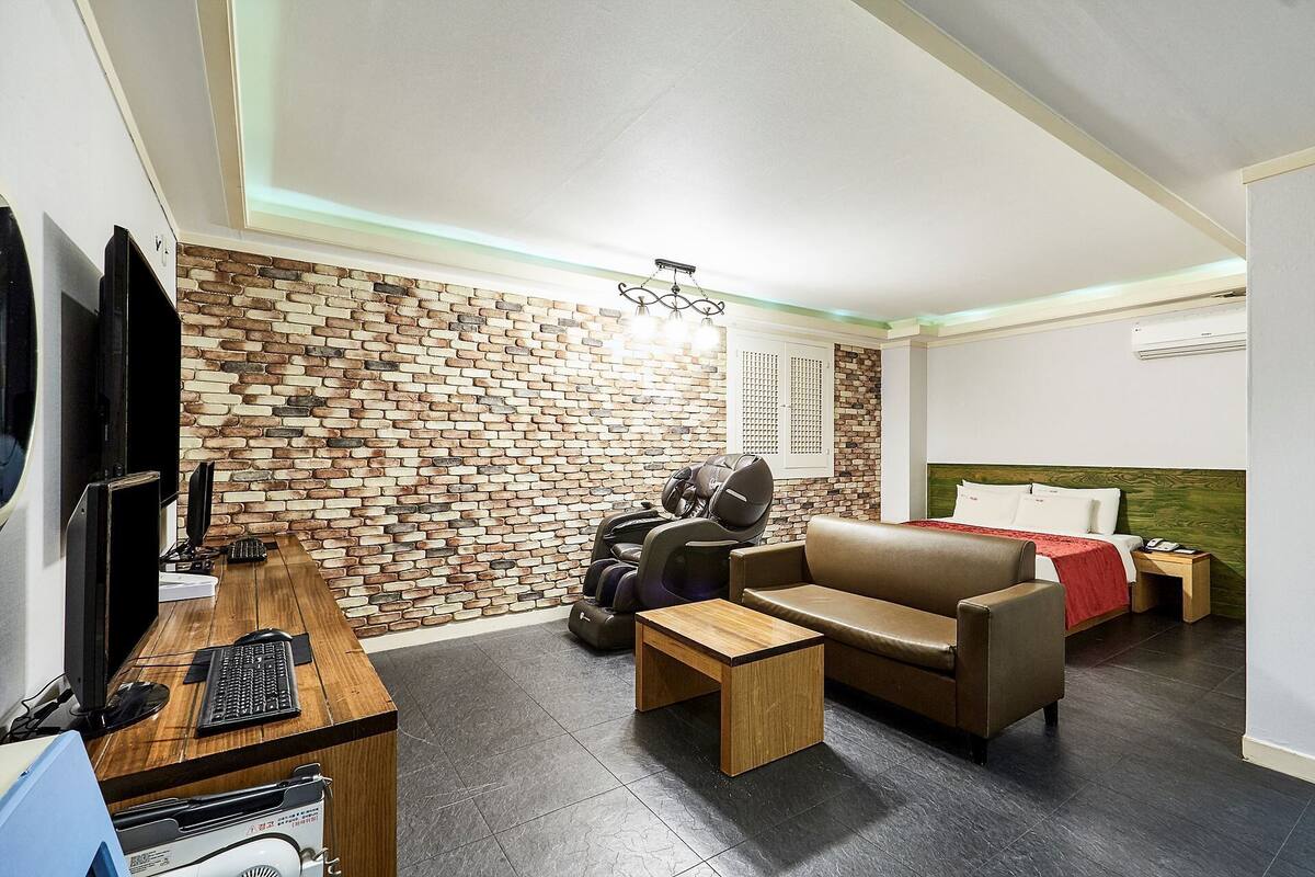 A compact hotel room with a brick-patterned wall, a sofa, a small wooden table, a massage chair, a TV, and a bed with a headboard.