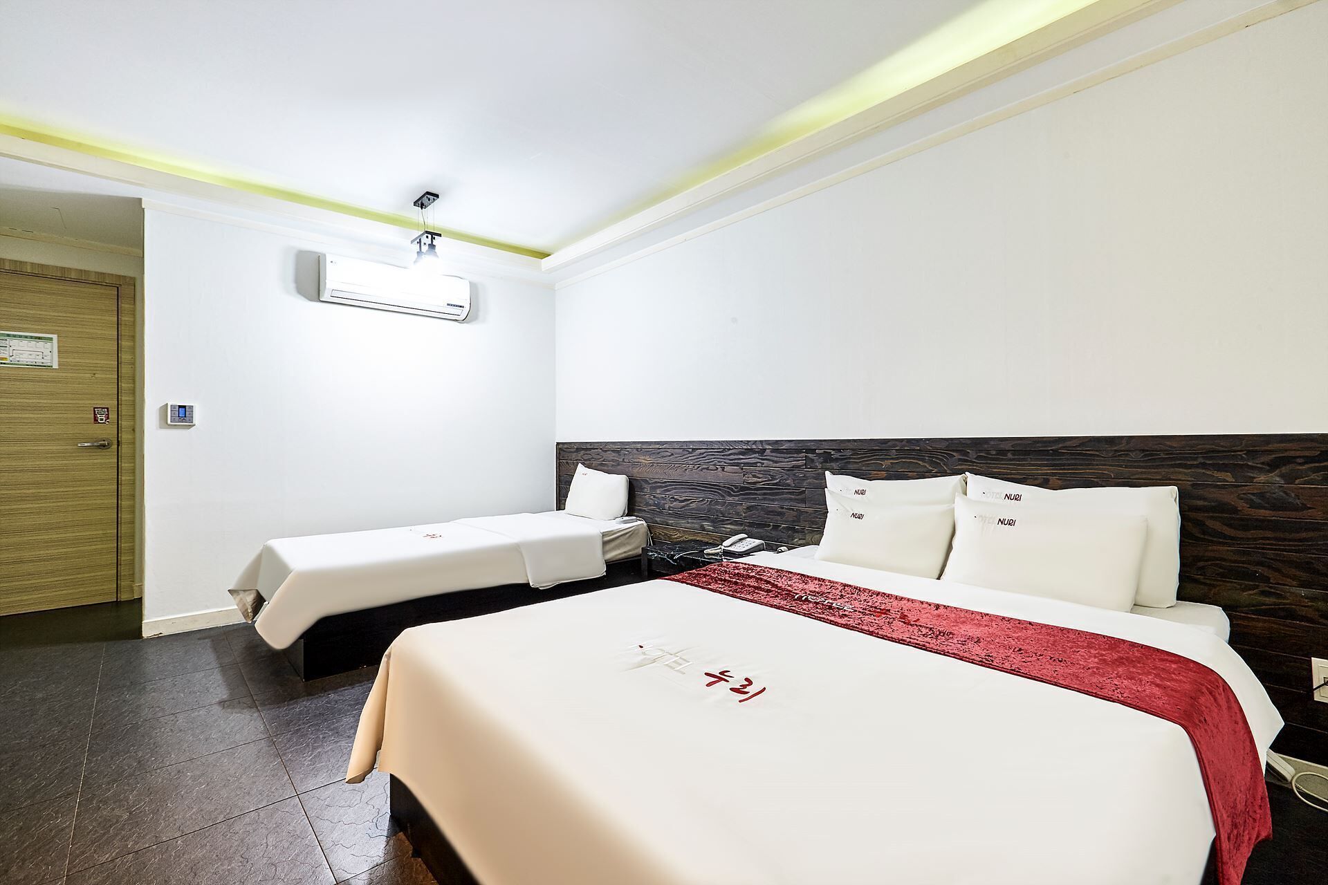 A hotel room with two beds, a wooden door, an air conditioning unit, and a red throw blanket.