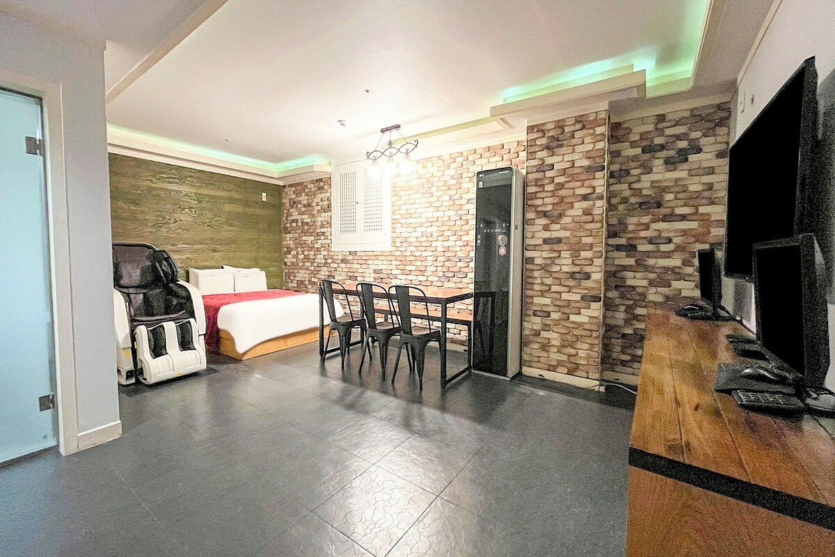 Una habitación de hotel moderna con cama, un área de comedor con sillas, un televisor y una pared de ladrillo.