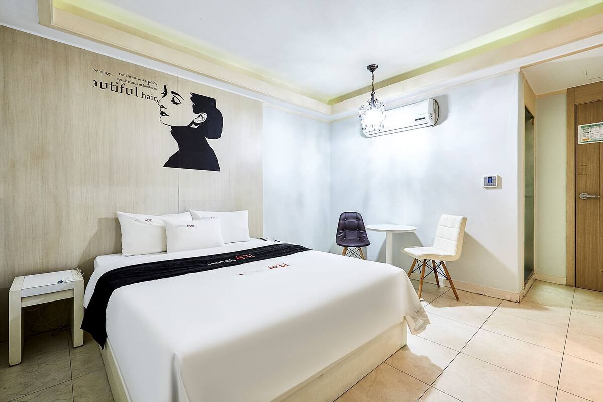 A hotel room with a large bed, a bedside table, a chair, a small table, and an air conditioning unit.