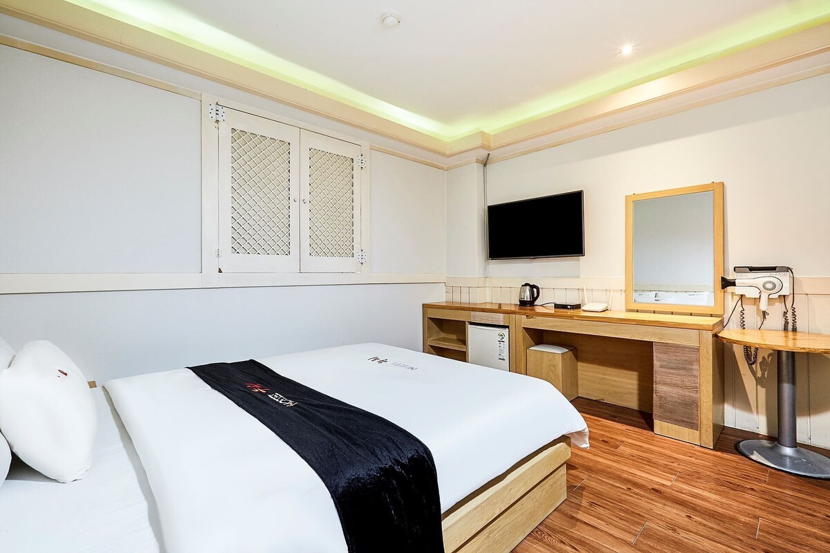Habitación de hotel con cama, escritorio con espejo, televisor y refrigerador pequeño.