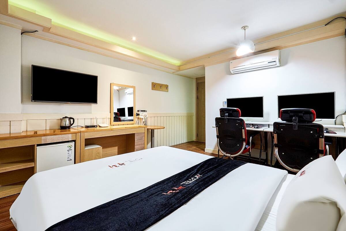 A hotel room with a bed, a desk with a computer, a chair, a TV, and an air conditioning unit.