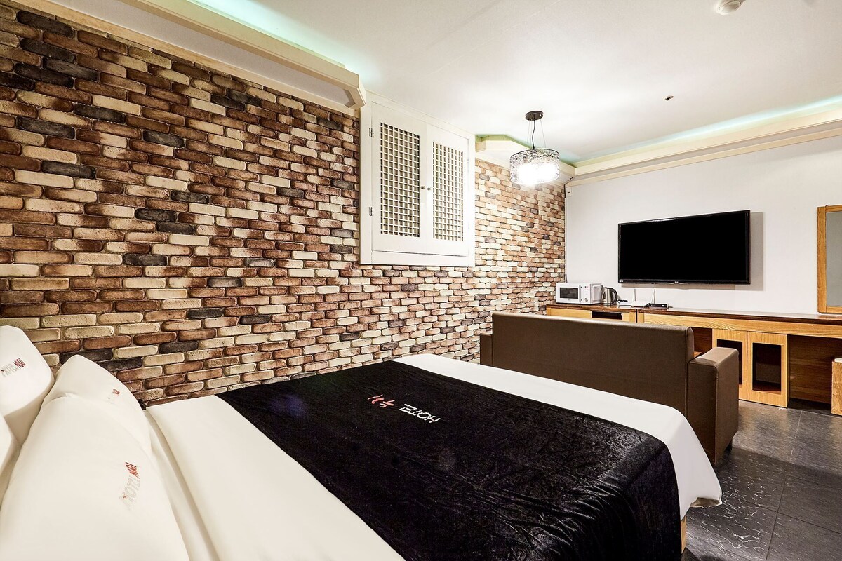 Habitación de hotel con pared de ladrillo, cama con cubrecama negro, televisión, escritorio con computadora y ventana con persianas.