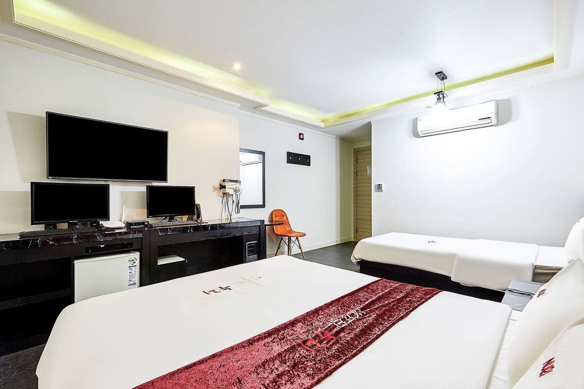 A hotel room with a bed, two bedside tables, a desk with a computer, a chair, a TV, and an air conditioning unit.