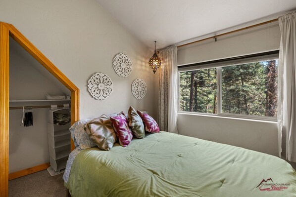 Een slaapkamer met een bed, een inloopkast, decoratieve wandkunst en een raam met uitzicht op een bos.