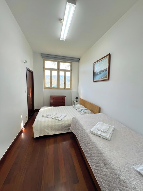 A hotel room with two beds, a painting on the wall, a window with a view, and a ceiling light.