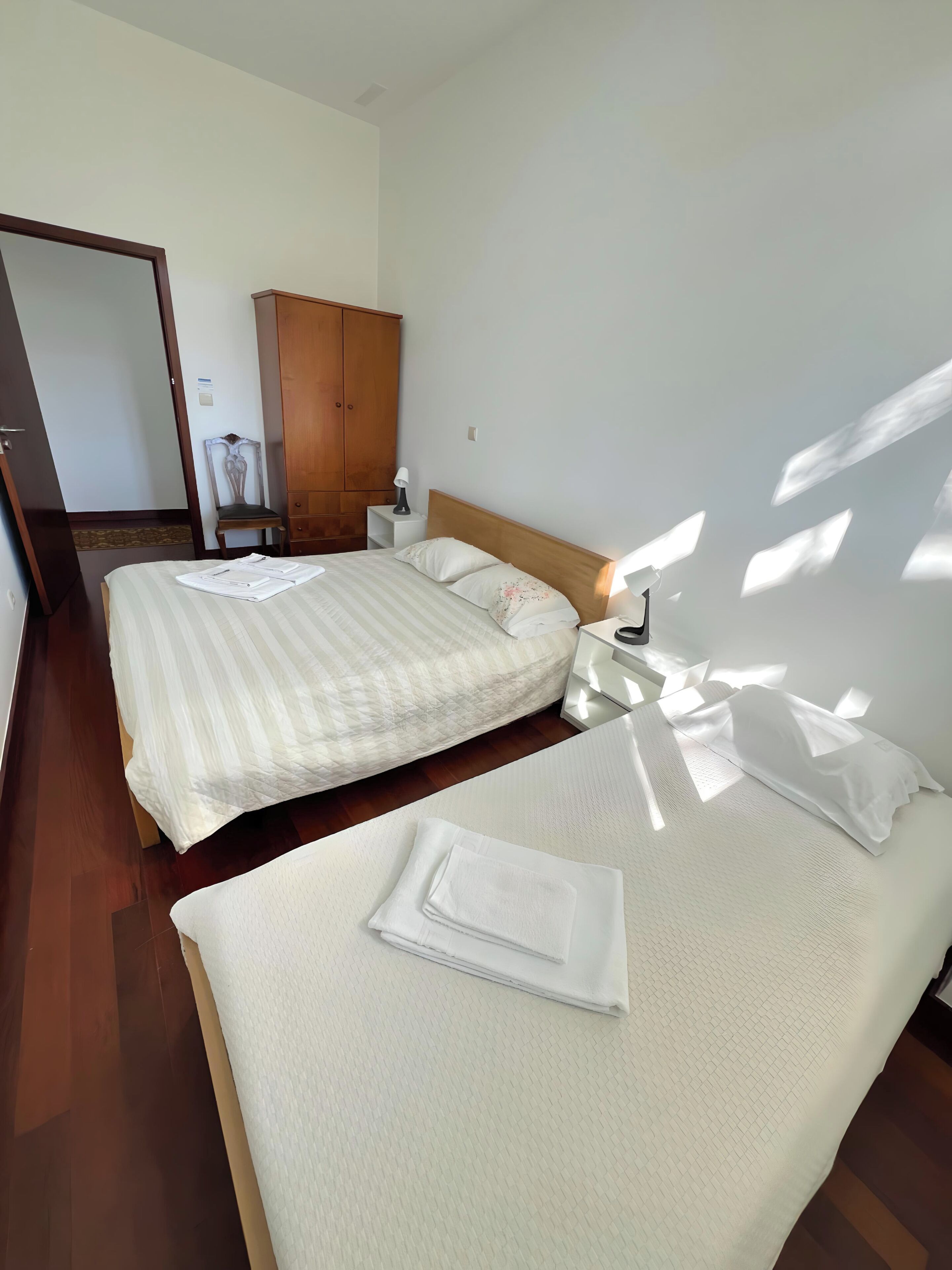 Una habitación de hotel con dos camas, un armario de madera, una mesita de noche con una lámpara y una silla.