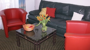 Un salon avec une table basse en verre, des fauteuils rouges, un canapé en cuir noir, un vase avec des fleurs et un tableau encadré au mur.