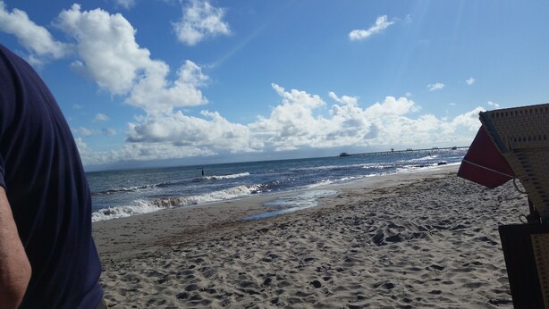 Une plage avec une jetée qui s’avance dans l’océan, une plage de sable et un ciel dégagé avec quelques nuages.