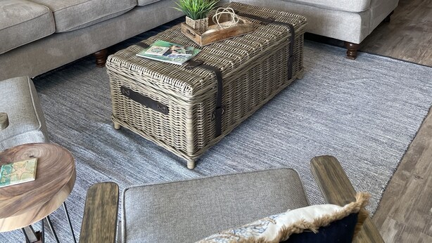 Un salon avec deux canapés gris, une table basse en osier, une petite table d’appoint et une grande plante en pot.