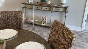 Un coin repas comprenant une table ronde, des chaises en osier, une console ornée d’objets décoratifs et un miroir.