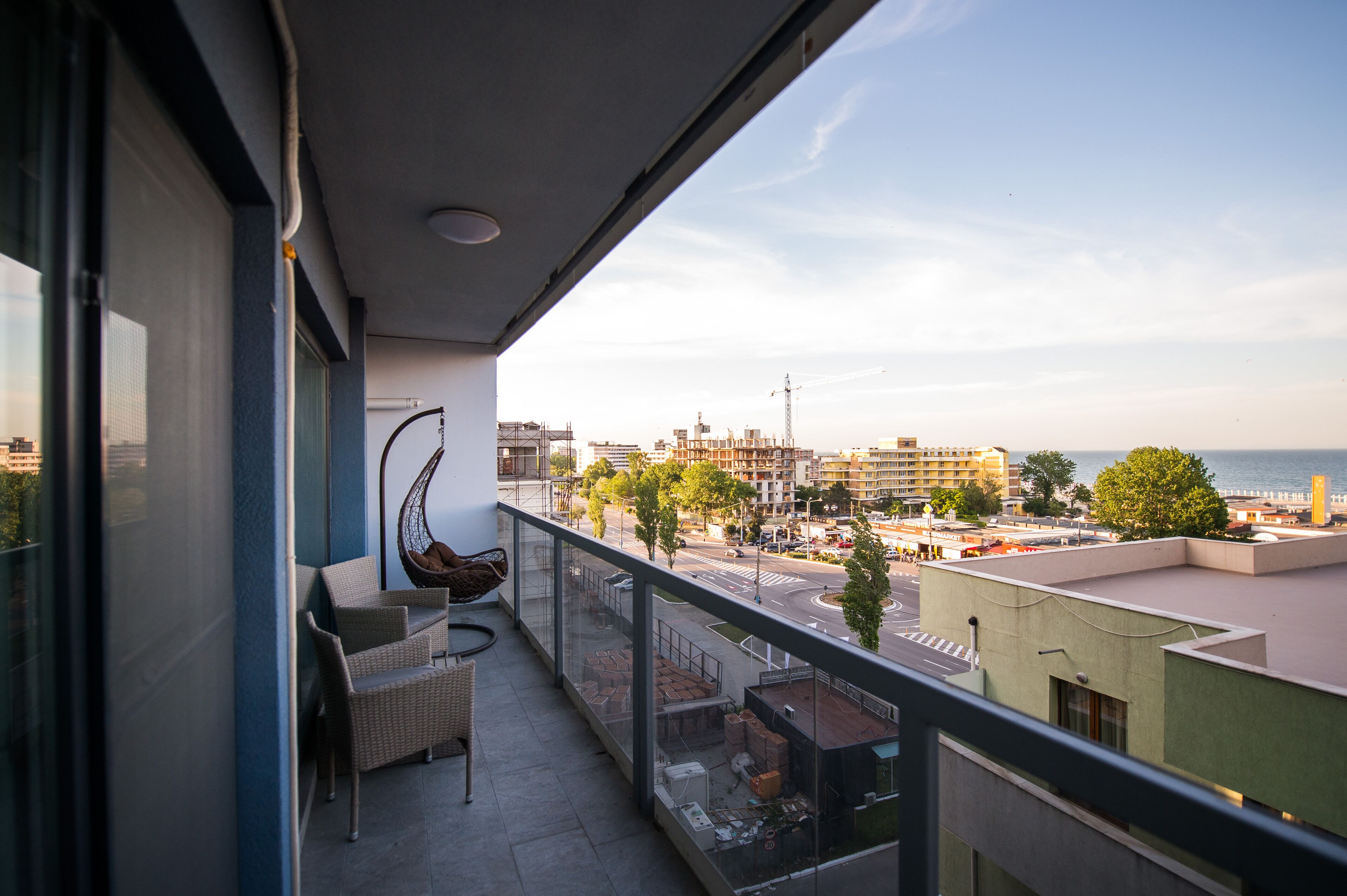 A balcony with wicker furniture, a hanging chair, and a view of a cityscape with buildings and a body of water.