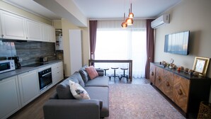 Ett modernt kök med vita skåp, en inbyggd ugn och en mikrovågsugn. Det finns en grå soffa med en prydnadskudde och ett soffbord med fyra stolar. Ett träskåp med dekorativa föremål står på höger sida.