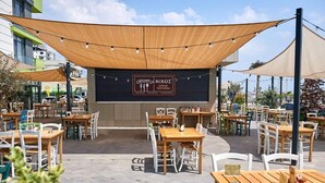 Ett utomhusområde med bord och stolar under ett skuggande tak, en skylt för den grekiska restaurangen Nikos, och utsikt över de omgivande byggnaderna.