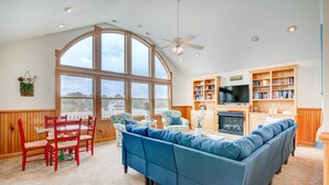A spacious living room with a large window, a fireplace, a television, and a bookshelf. - 1069 - Seadation - Lakeview Corolla Escape w/ Pool & Hot Tub (Corolla)