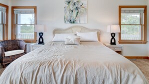A spacious bedroom with a large bed, two nightstands, a wicker chair, and a painting above the bed. - 1069 - Seadation - Lakeview Corolla Escape w/ Pool & Hot Tub (Corolla)