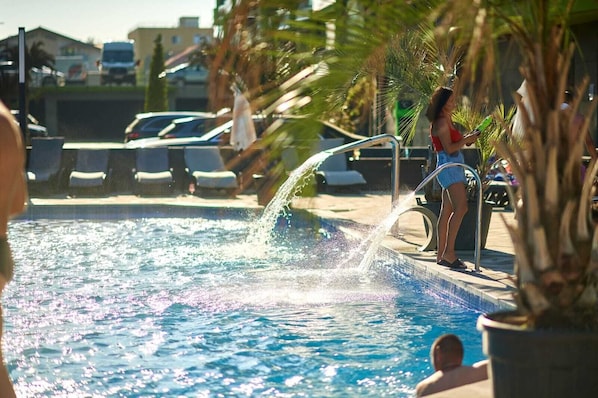A swimming pool with a water fountain, surrounded by palm trees and lounge chairs.