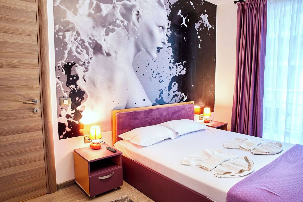 A hotel room with a bed, nightstands, a wall mural, and a window with curtains.