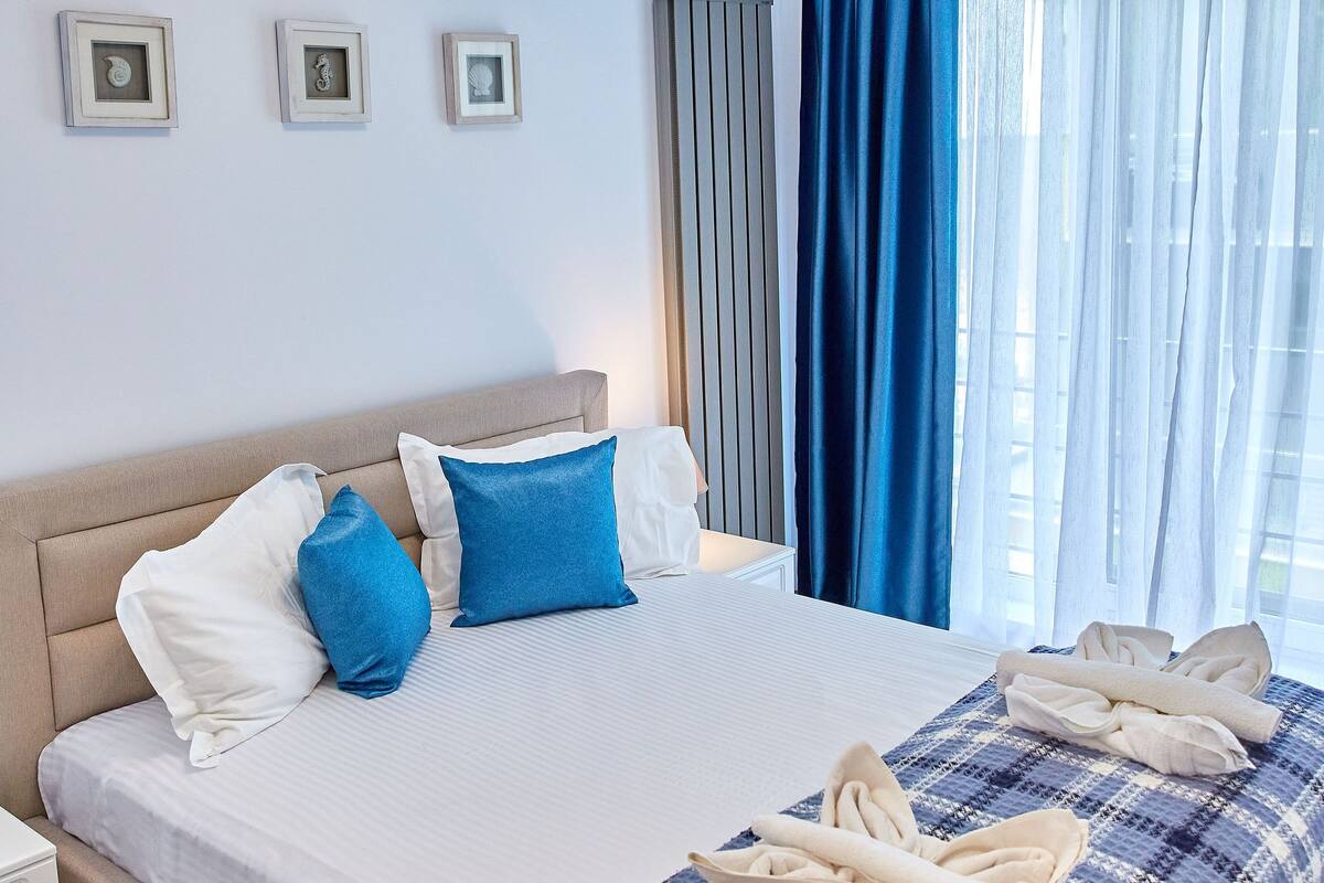 A neatly made bed with a plaid blanket, a bedside table, and a window with blue curtains.