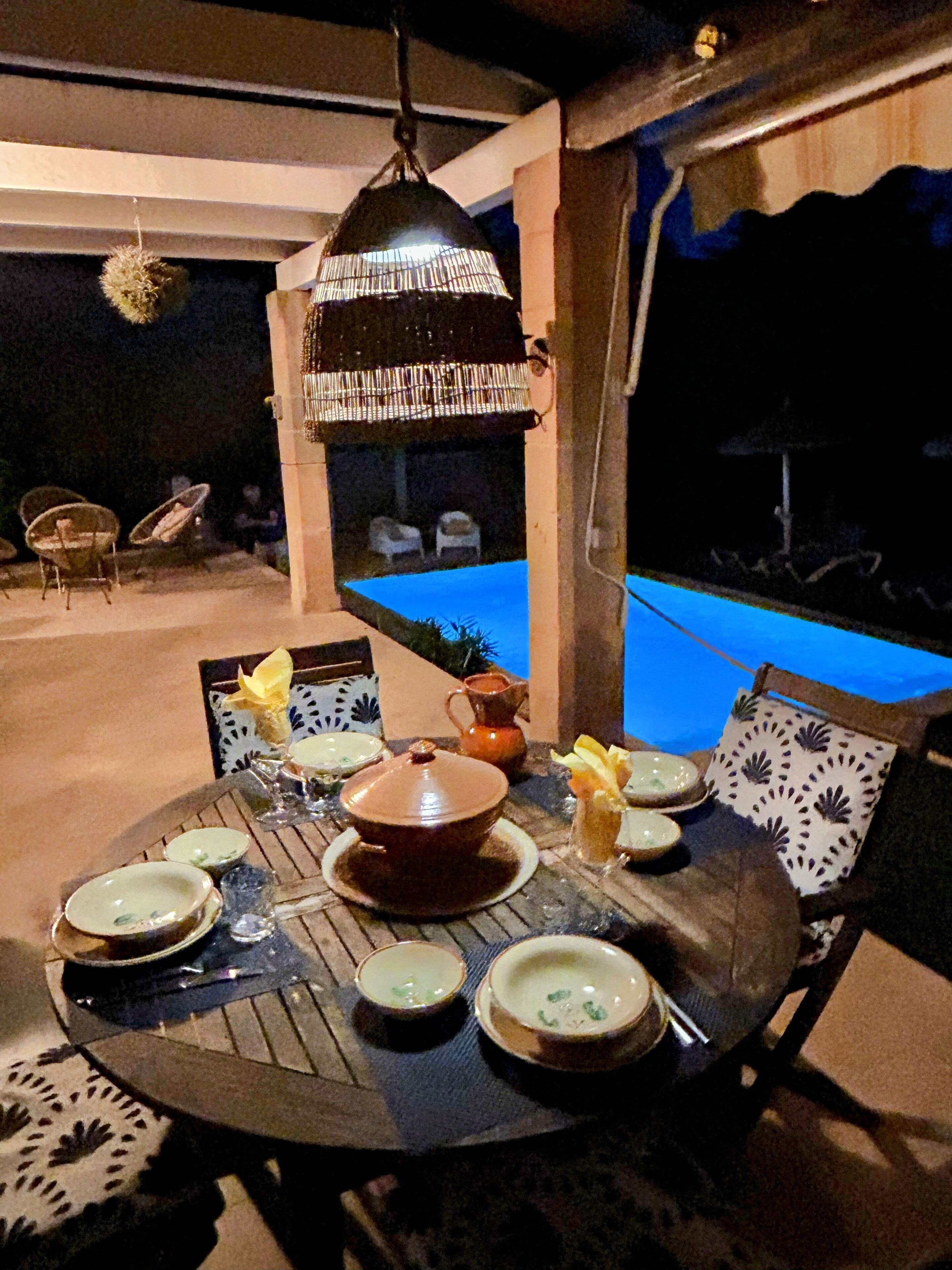 An outdoor dining area with a round table set for four, surrounded by chairs. A hanging light fixture above the table and a pool in the background are visible.