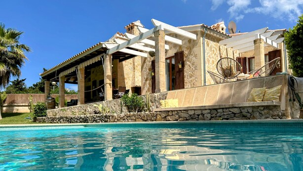 Zona de piscina con muro de piedra, pérgola y vista a un edificio con antena parabólica.