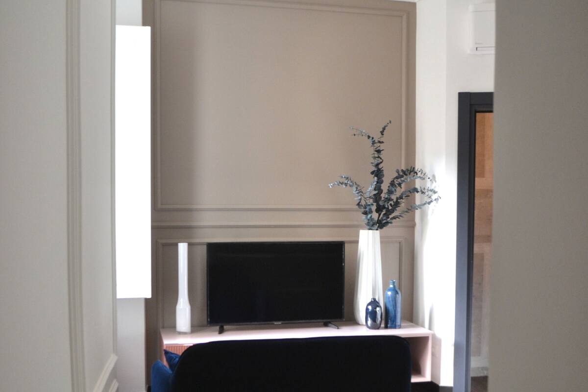 Un salón moderno con un sofá de color oscuro, un televisor de pantalla plana y un jarrón decorativo con una planta.