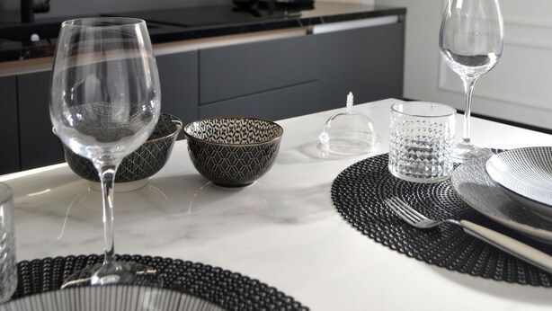 A modern kitchen with dark cabinetry, a countertop, and a dining area set with a white tablecloth, black placemats, and clear glassware.