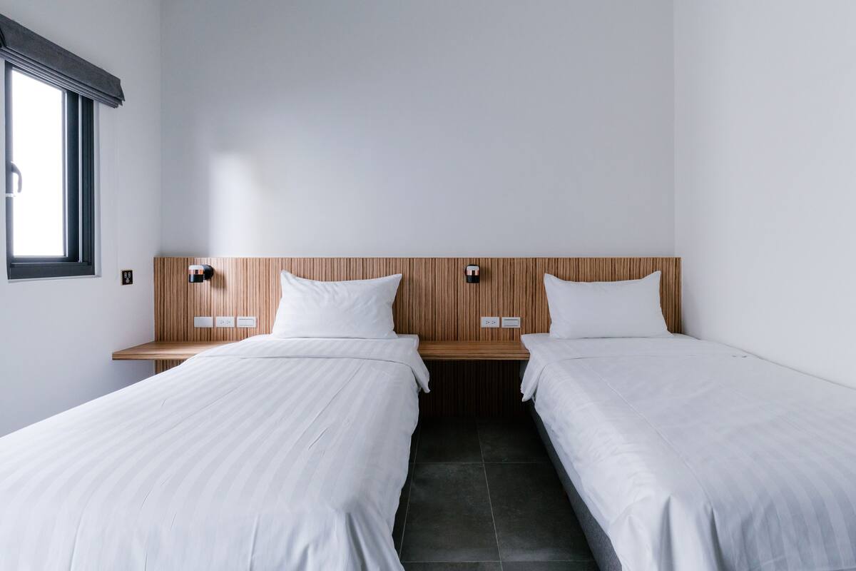 A hotel room with two single beds, wooden headboards, bedside tables, and a window with blinds.