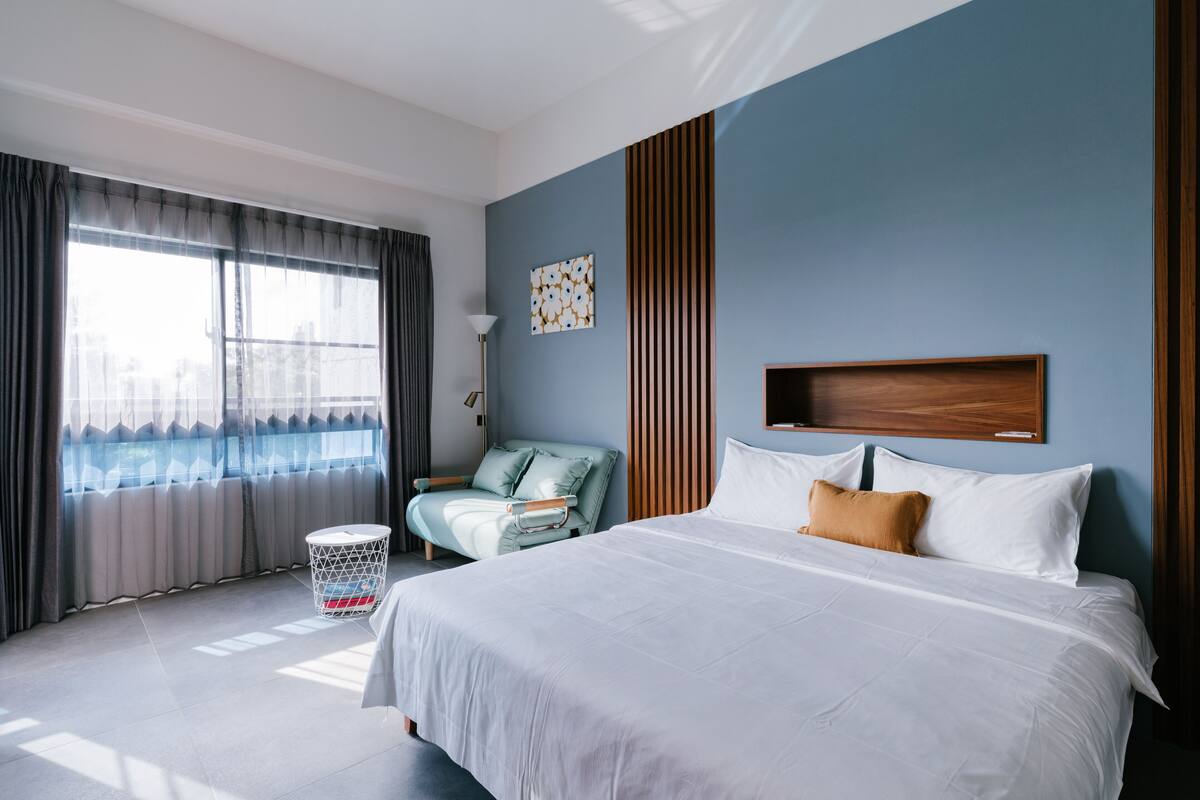A modern hotel room with a large bed, a seating area with a chair, a small table, and a window with curtains.