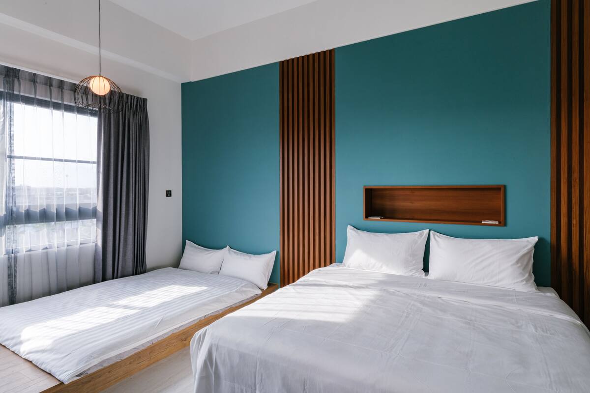 A hotel room with two beds, a wooden headboard, teal walls, and a window with curtains.