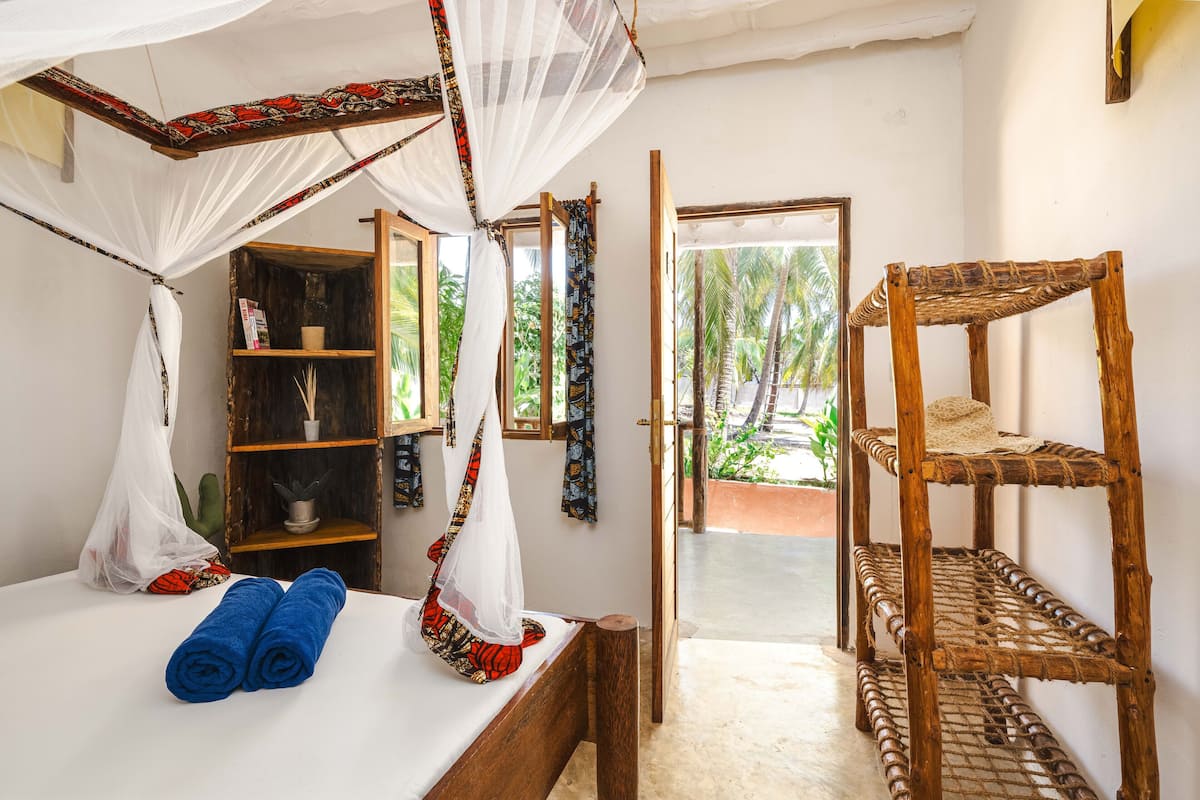 Quarto com cama, prateleira de madeira e janela com vista para palmeiras.