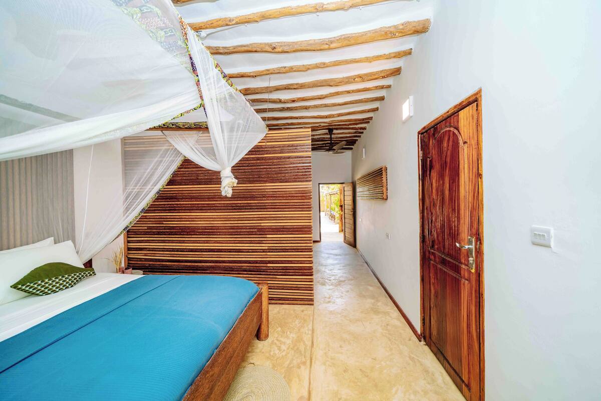 Quarto com cama, ripas de madeira, porta de madeira e teto com vigas aparentes.