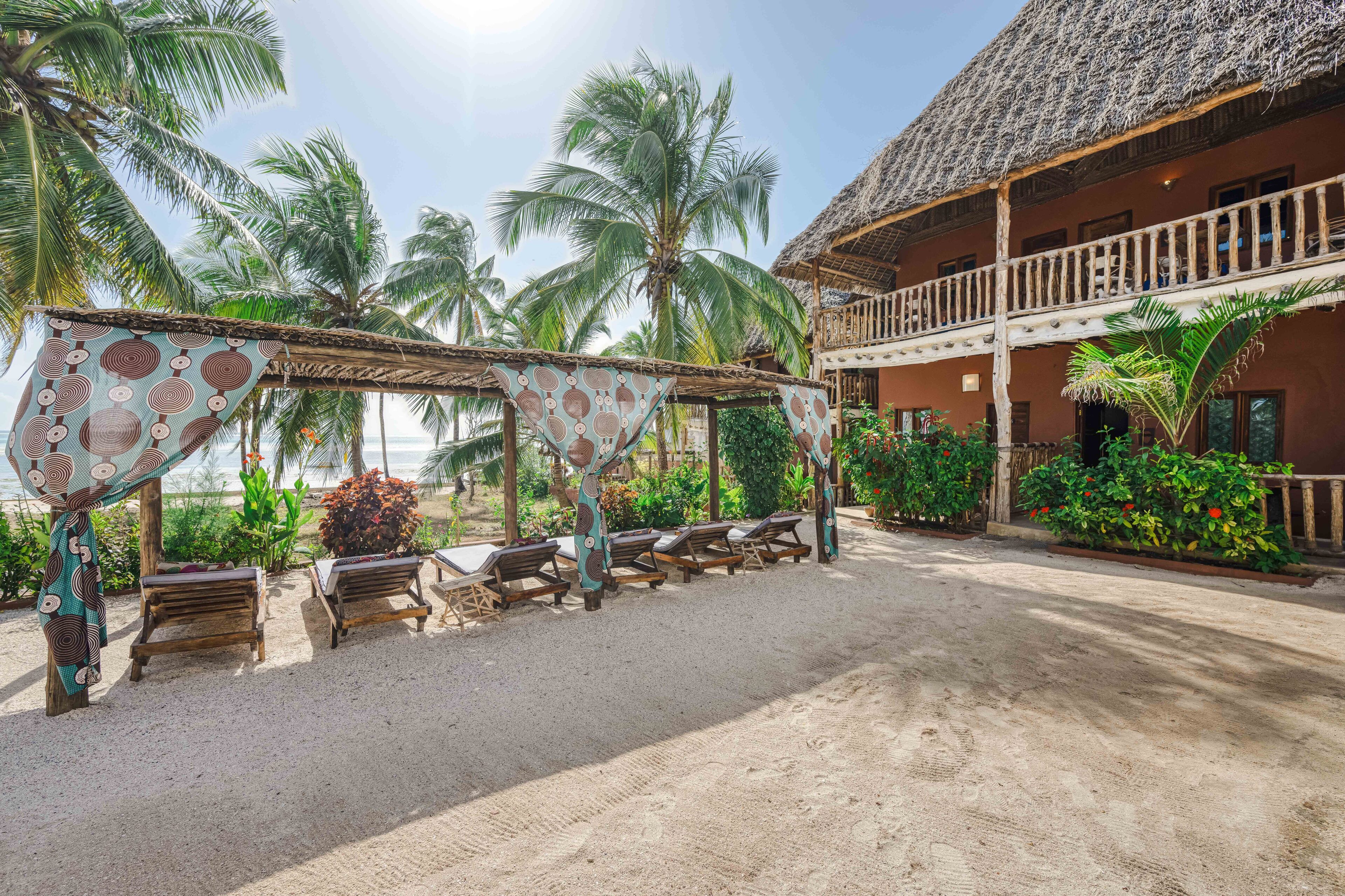 A beachside resort with thatched-roof buildings, wooden lounge chairs, and palm trees.