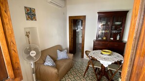 A living room with a brown sofa, a standing fan, a patterned floor, a wooden cabinet, and a dining table with a white tablecloth. - Traditional Eolian House "Luna"- Unique experience (Lipari)