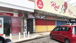 Vista de la calle con un coche rojo estacionado en la banquina, un restaurante de comida rápida abierto las 24 horas y una tienda cerrada con un toldo rojo.