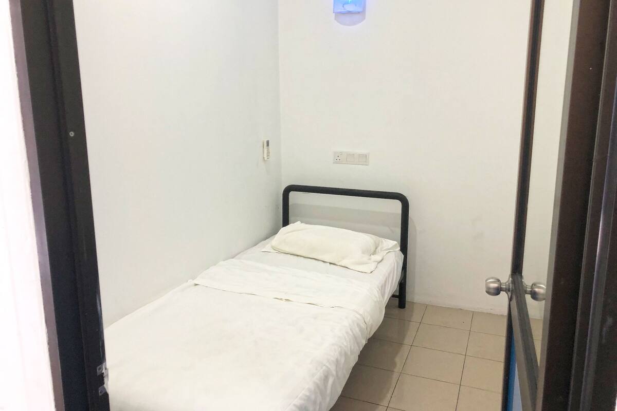 A small, single-bed room with a white bedspread, a black metal headboard, and a wall-mounted light fixture.