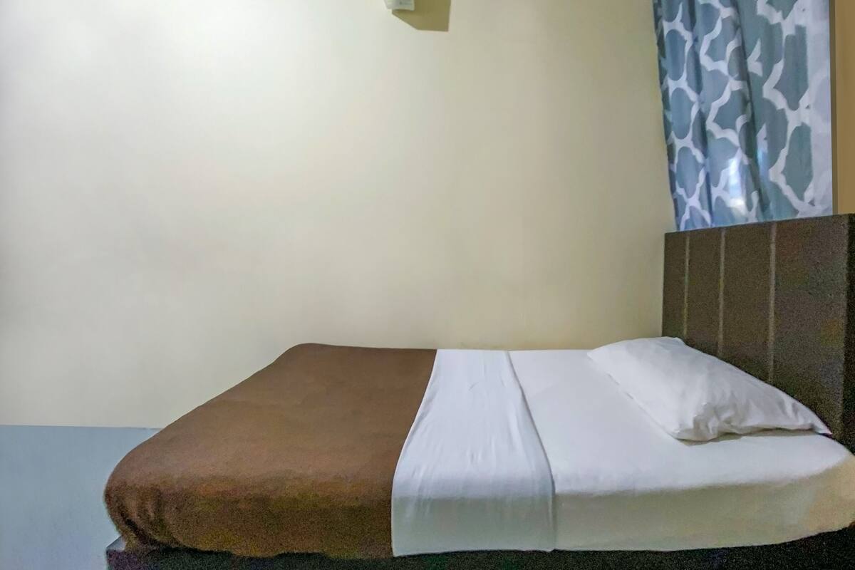Ein ordentlich bezogenes Bett mit brauner Decke und weißen Laken, ein Kopfteil mit strukturierter Musterung und eine an der Wand befestigte Leuchte.