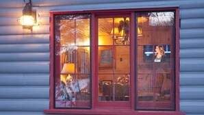 Une cabane en rondins dotée d’une grande fenêtre, d’une lampe suspendue et d’un intérieur visible, avec une chaise et une table.